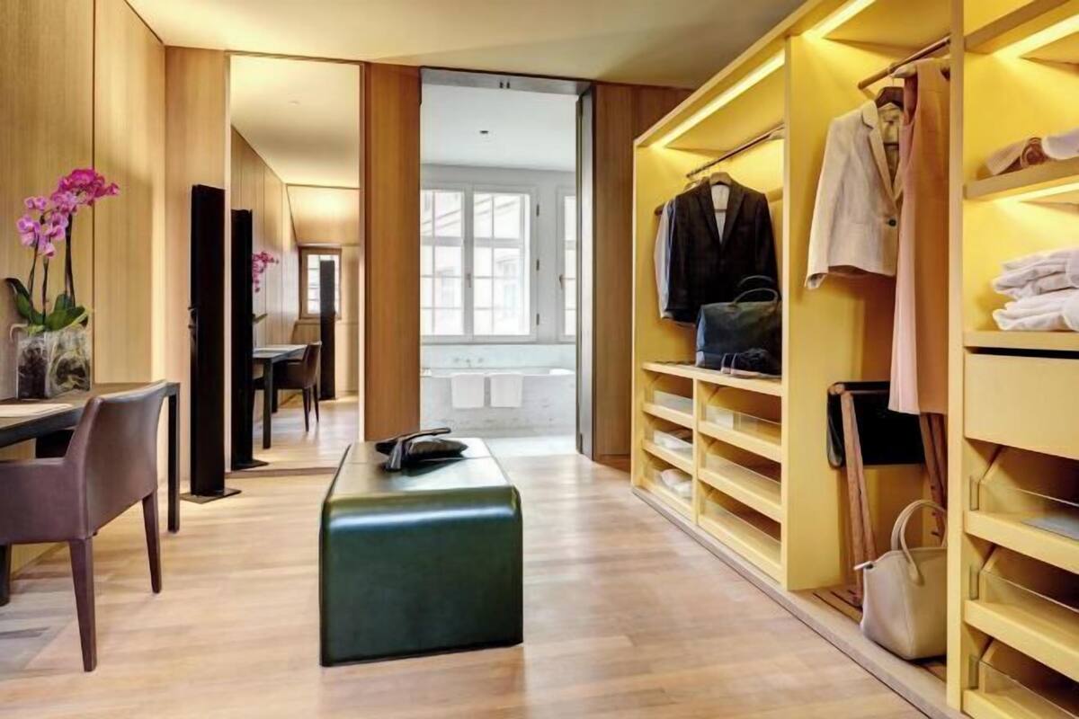 A modern hotel room with a wooden floor, a desk with a chair, a bench, a walk-in closet with shelves, and a view of the outside.