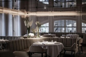 Un restaurant avec des tables rondes et des banquettes, de grandes fenêtres et un plafond haut.