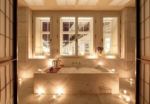 A spacious bathroom with a large bathtub, a window with a view of a building, and candles on the floor. - Hotel Cafe Royal (London)