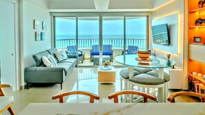 A modern living room with a glass dining table, grey sofa, and blue chairs, overlooking a balcony with a view of the sea.