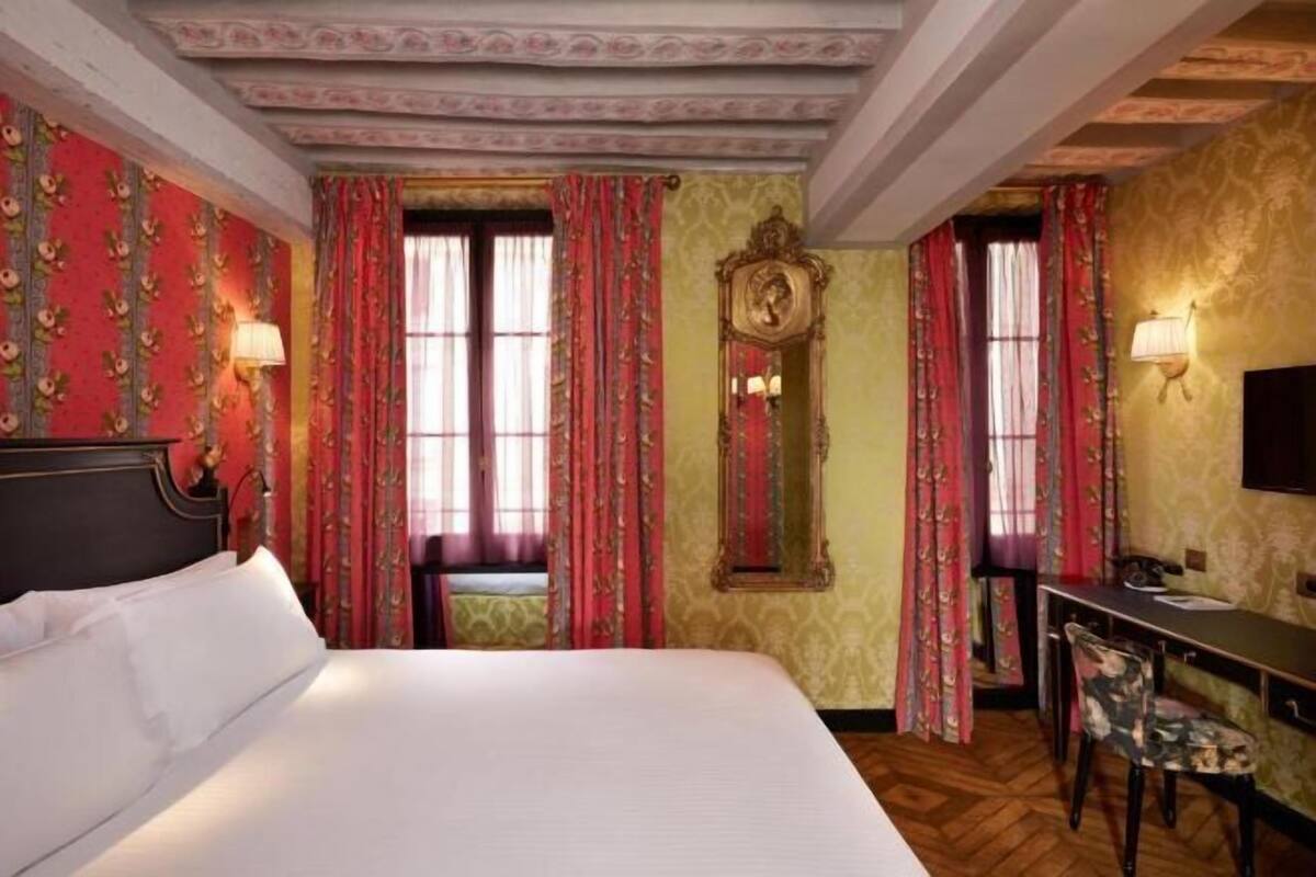 A hotel room with a large bed, a desk with a chair, and two windows with curtains.