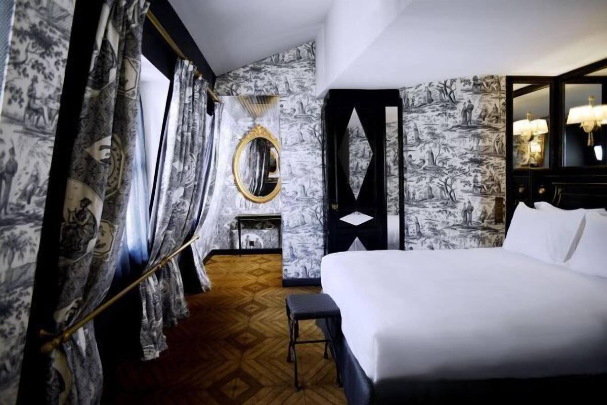 A hotel room with a large bed, a wooden floor, patterned wallpaper, a mirror, and a small table.