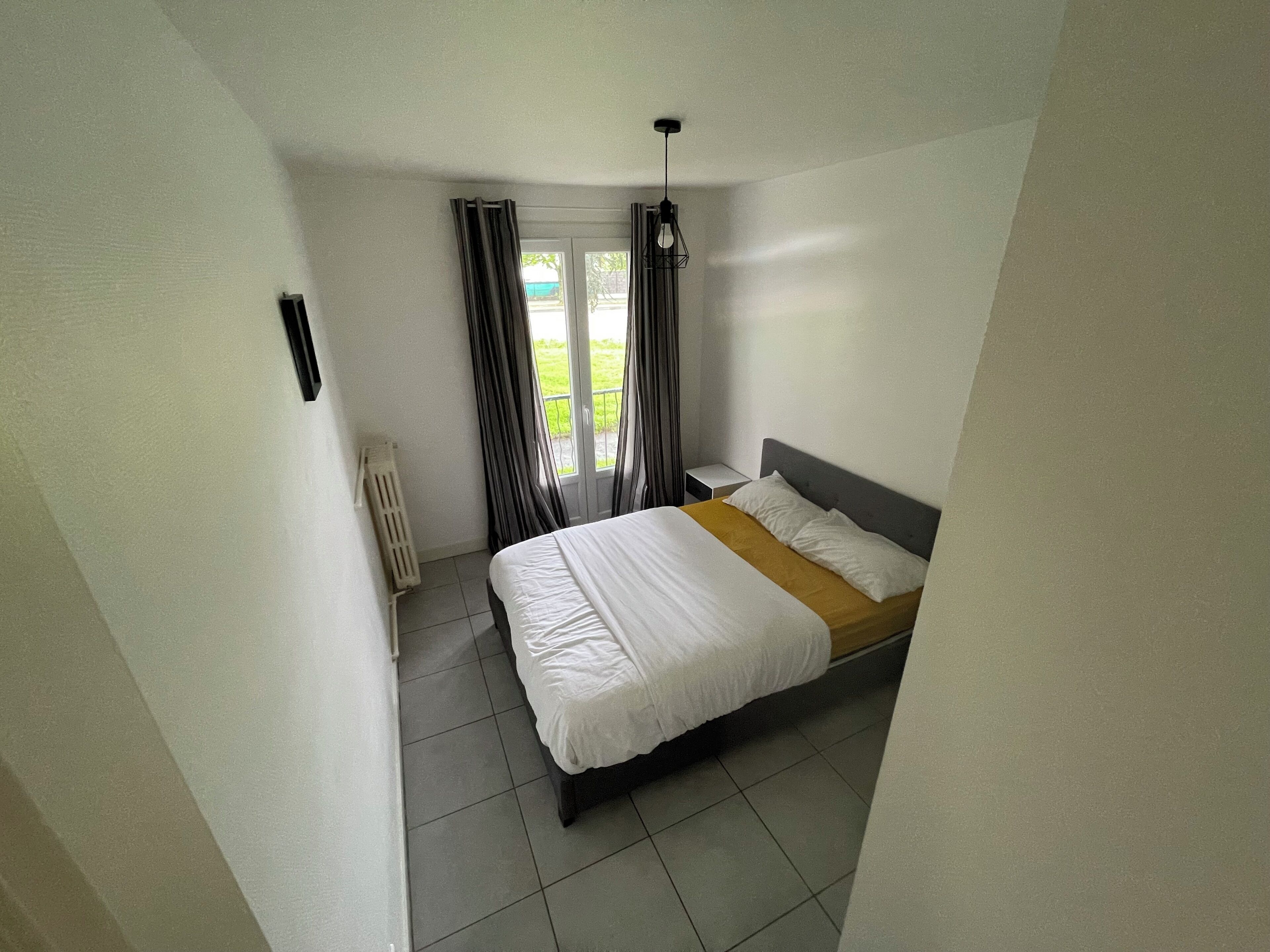 A bedroom with a bed, a radiator, a window with curtains, and a view of greenery outside.