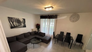 Ein Wohnzimmer mit Ecksofa, Couchtisch, Esstisch mit Stühlen, einer Wanduhr und einem Heizkörper mit Blumen.