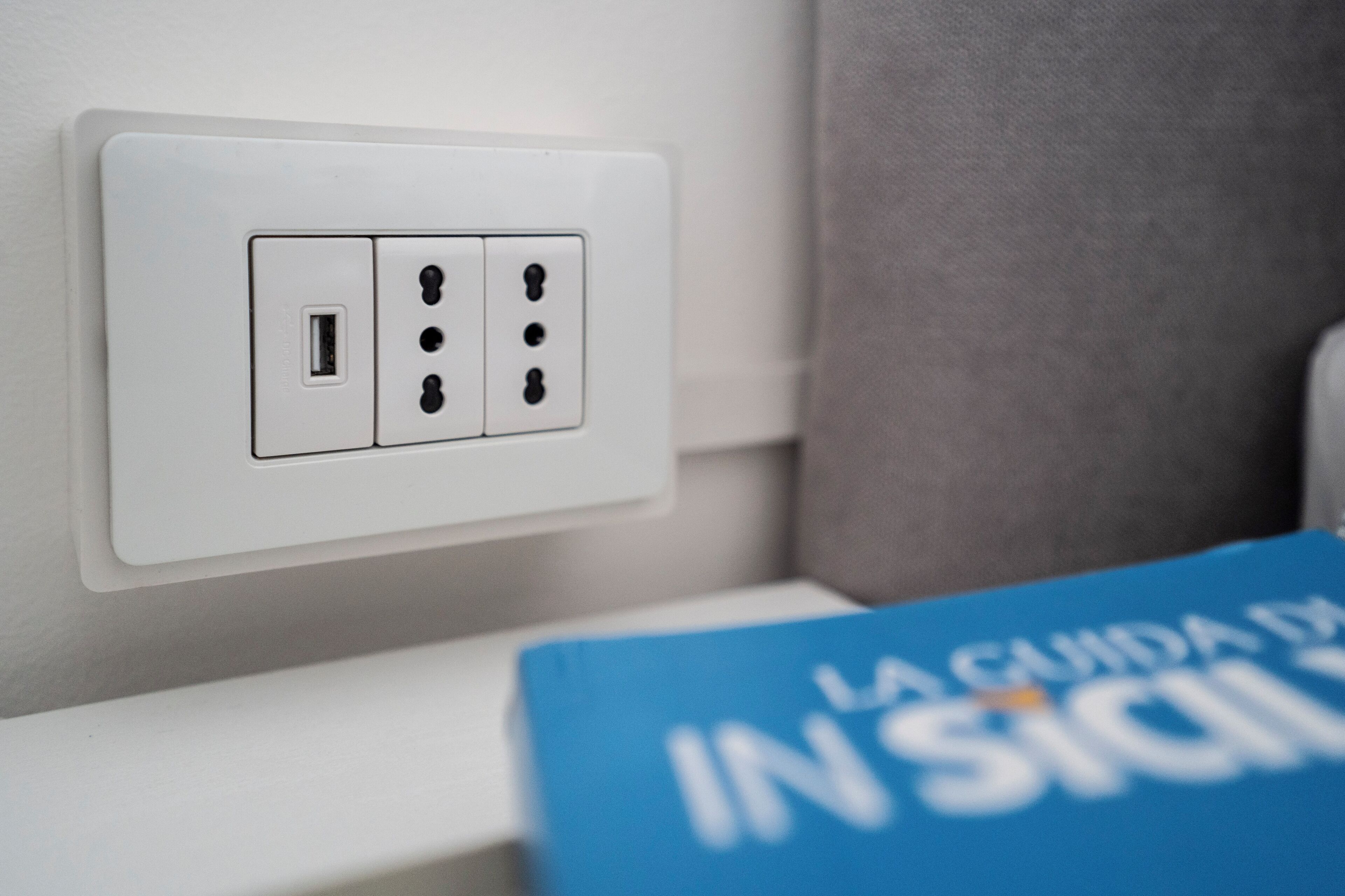 Une prise électrique blanche dotée d’un port USB et de trois prises standard, située à côté d’un rideau gris.