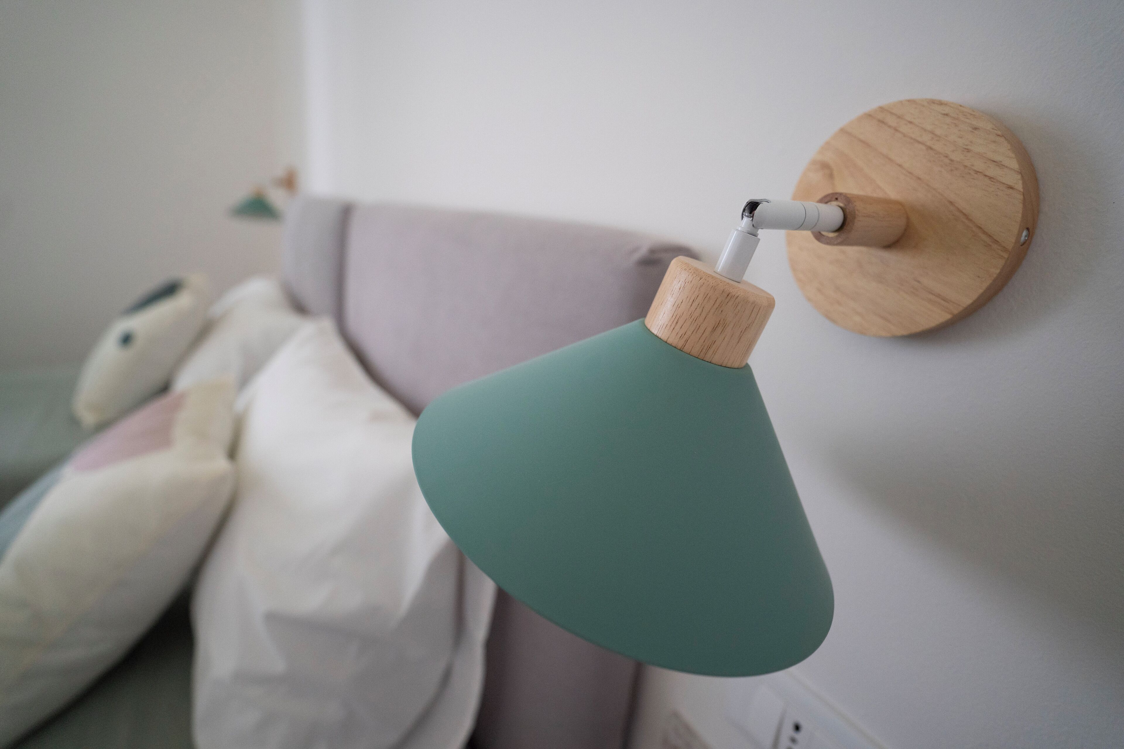 Une lampe murale avec un pied en bois et un abat-jour vert, un lit avec des oreillers et un mur blanc.
