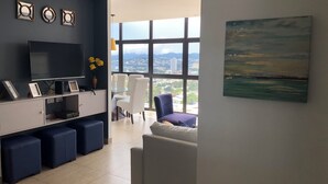 Un salon moderne doté d’un grand téléviseur, d’un canapé blanc, d’un fauteuil bleu et offrant une vue sur la ville grâce à de grandes fenêtres.