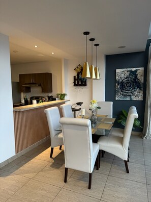 A modern dining area with a glass table and white chairs, a kitchen in the background, and a dark accent wall with a large abstract painting. - Best Location San Salvador, Escalón, Torre 91. N6 (San Salvador)