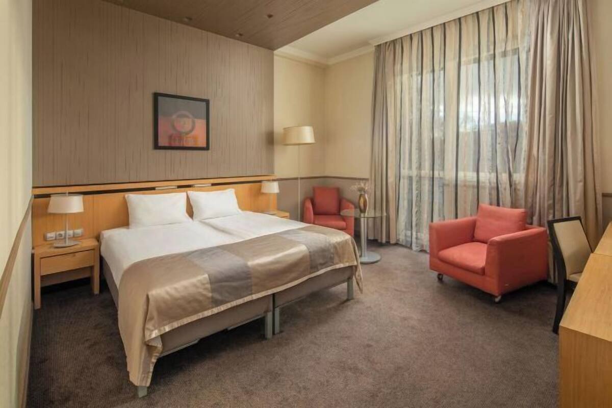 Une chambre d’hôtel comprenant un lit, deux tables de chevet, une chaise, un bureau et une grande fenêtre avec des rideaux.