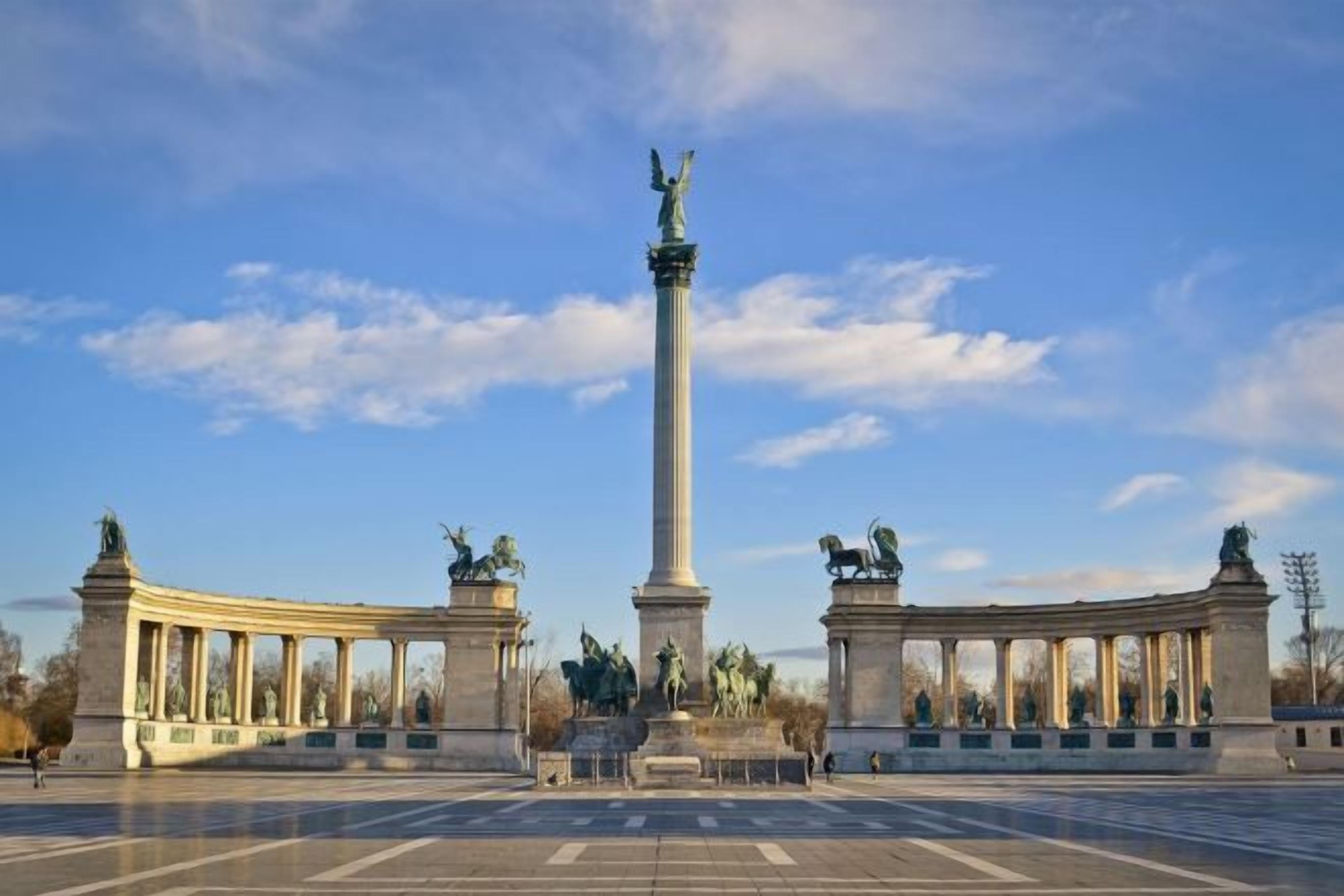 Uma grande praça aberta com um monumento central, ladeada por colunas e estátuas clássicas.