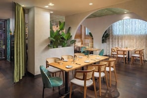 A restaurant with a long wooden table and chairs, a green curtain, and a plant. - Mamaison Hotel Andrassy Budapest (Budapest)