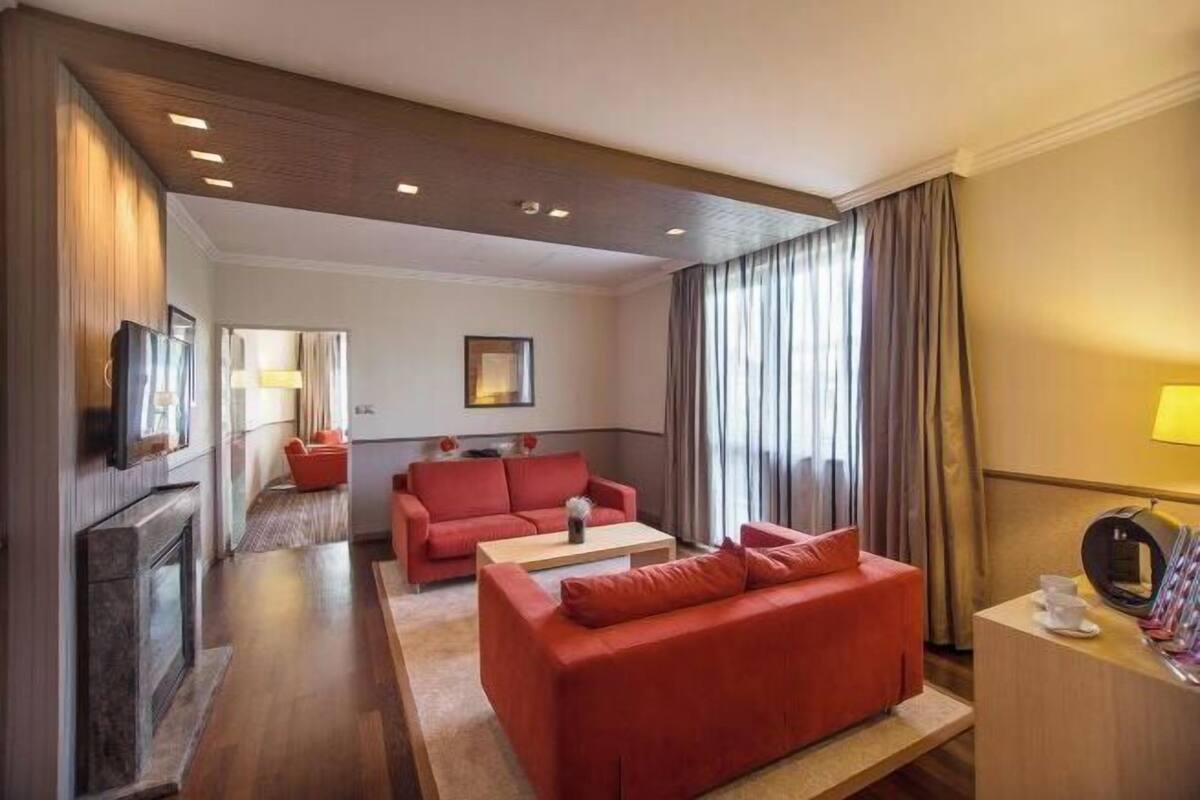 Une chambre d’hôtel avec un canapé rouge, une table basse en bois, une télévision et un bureau avec une lampe.