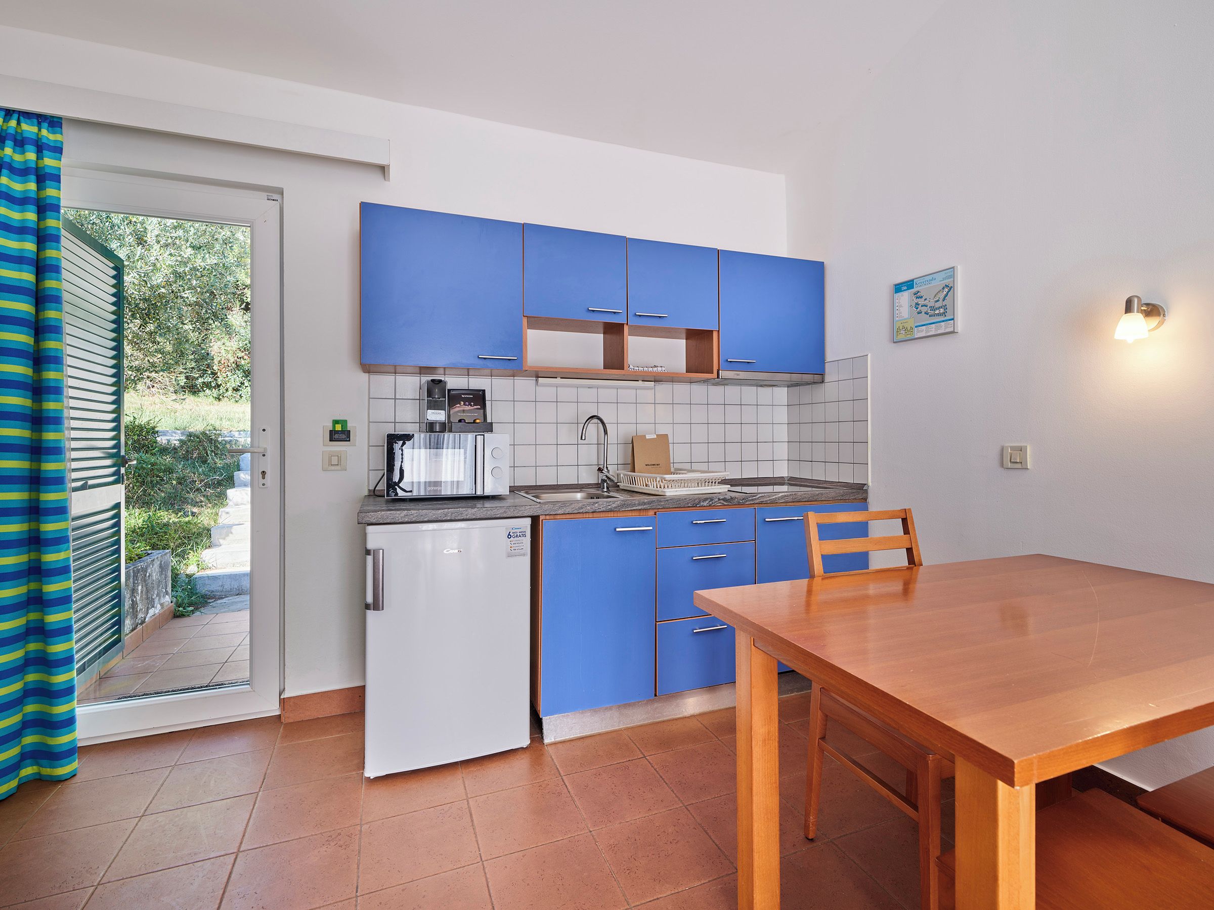 A compact kitchen with blue cabinets, a white refrigerator, a wooden dining table, and a view through the door.