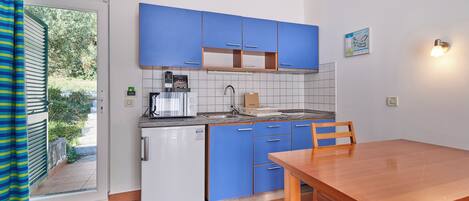 A compact kitchen with blue cabinets, a white refrigerator, a wooden dining table, and a view through the door.