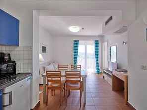 A compact kitchen with a refrigerator, stove, and microwave. There is a dining area with a table and chairs. A sofa is placed against the wall, and a window with curtains is visible. - Maistra Select Naturist Park Koversada Apartments (Vrsar)