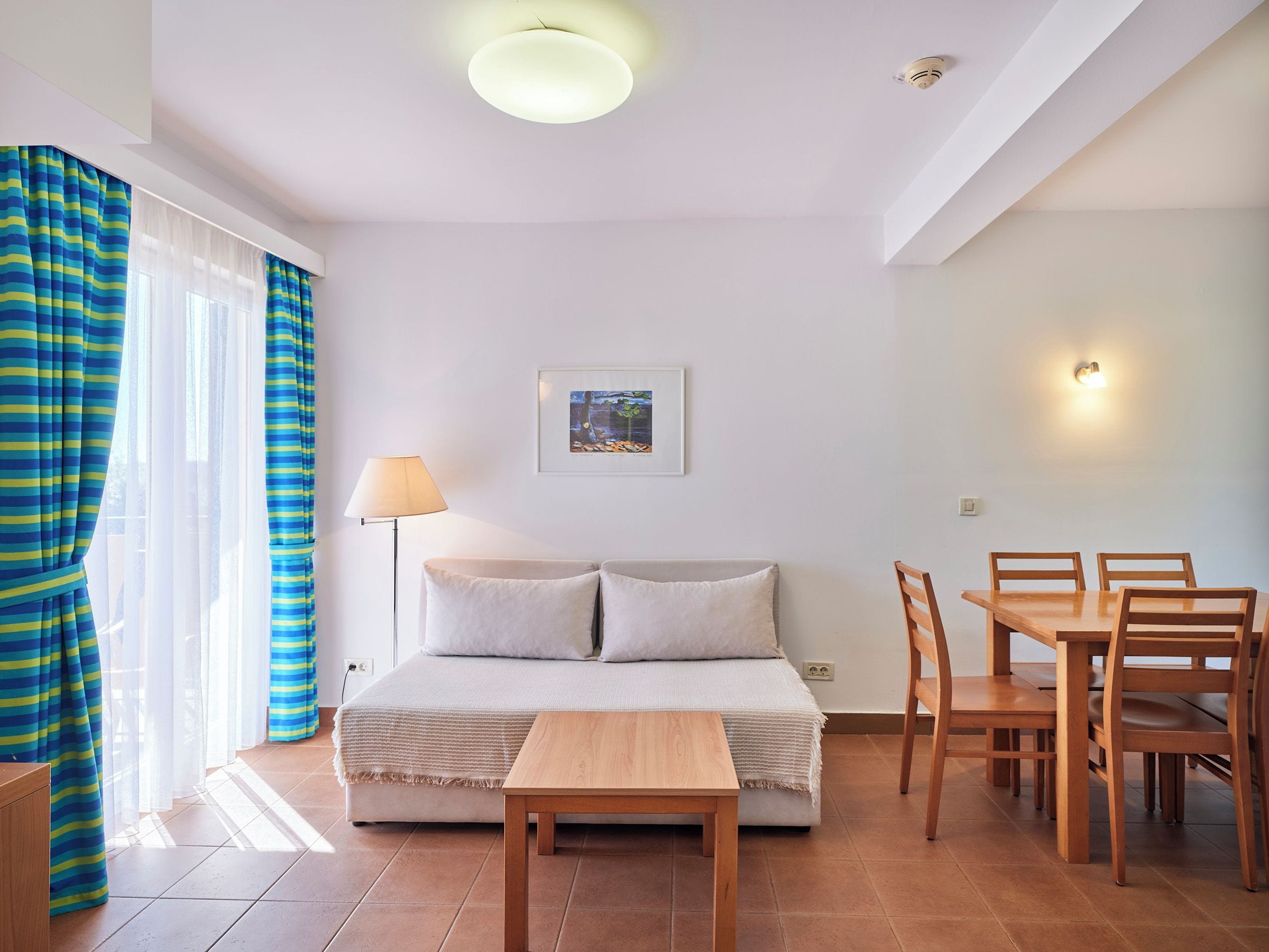 A bedroom with a bed, bedside table, lamp, and a dining area with chairs and a table.