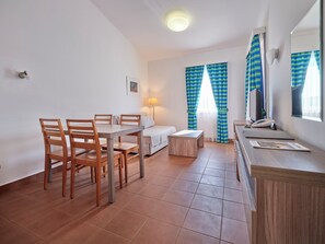 Il s’agit d’une pièce de taille modeste, comprenant une table à manger, des chaises, un canapé, une petite table de chevet et un bureau avec une chaise. On y trouve une lampe, un téléphone et un ordinateur sur le bureau. La pièce est dotée d’une fenêtre avec des rideaux et d’un luminaire au plafond.
