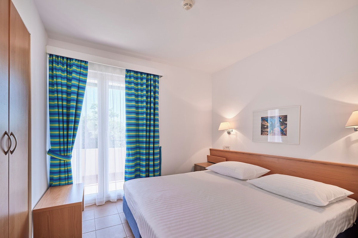 Habitación de hotel con cama, mesita de noche, lámpara, un cuadro en la pared y cortinas.