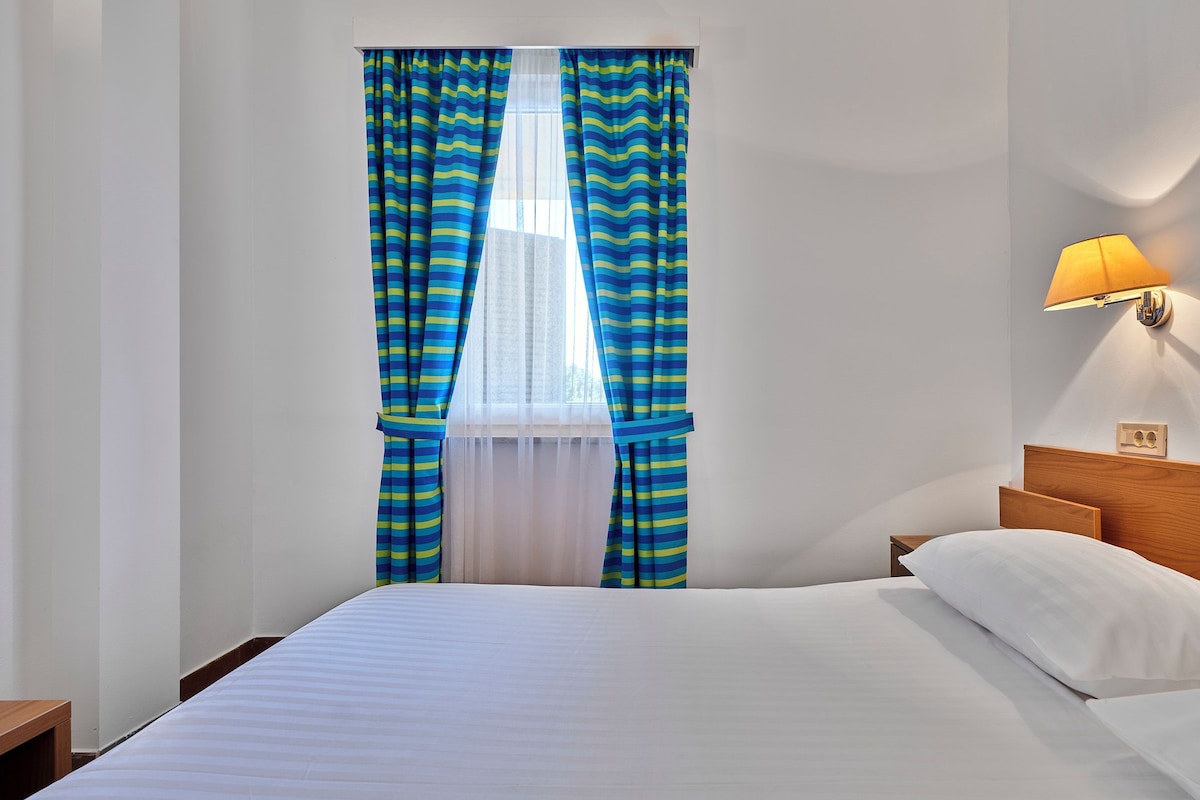 Una habitación de hotel con cama, cortinas, mesita de noche y lámpara de pared.