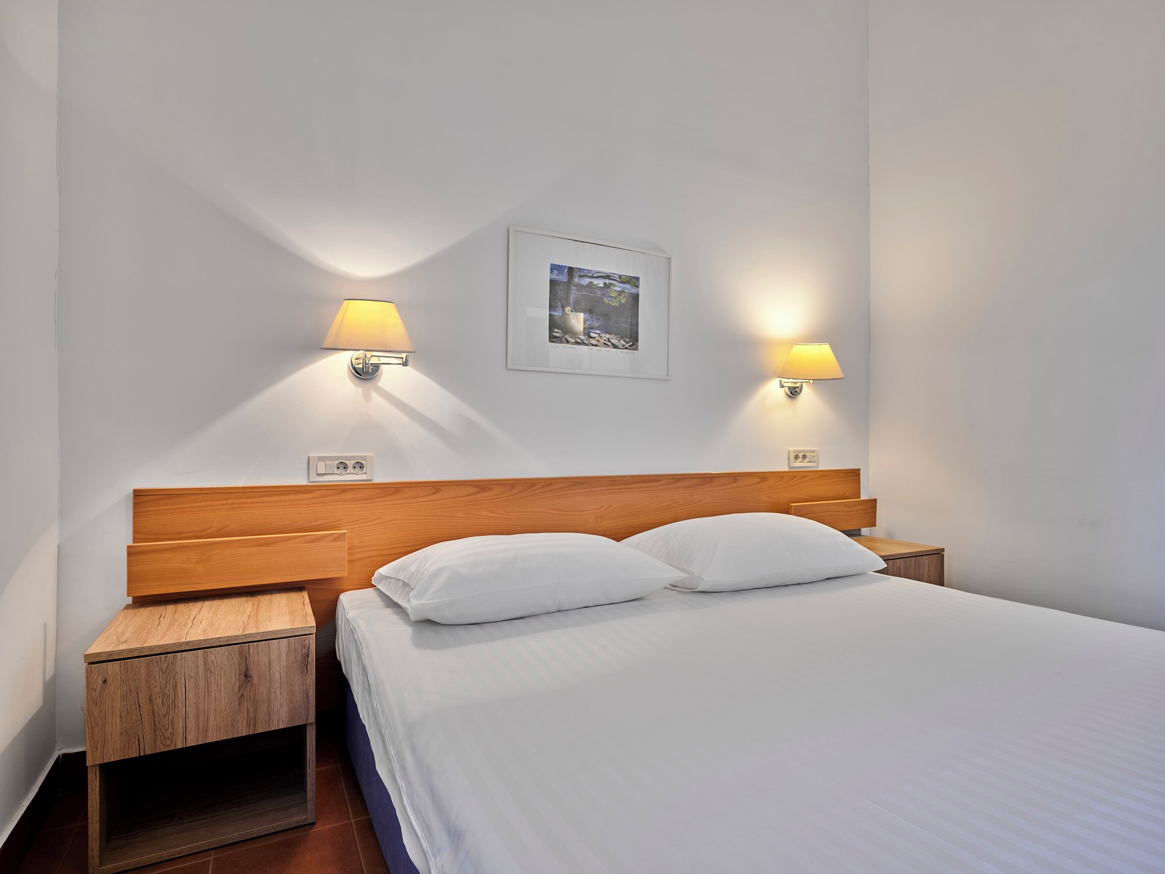 A neatly made bed with white linens and two pillows, flanked by wooden nightstands with lamps.