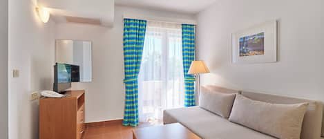 Une pièce comprenant un canapé, une petite table, un téléviseur, un téléphone, une lampe et une fenêtre avec des rideaux.