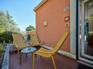 Een terras met gele rieten stoelen en een kleine tafel, een deur met een nummerbord, en uitzicht op groen.