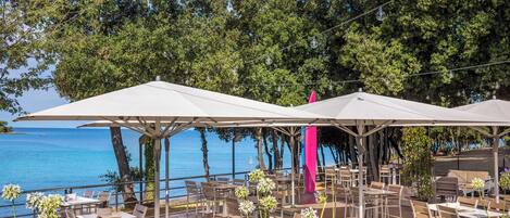 An outdoor dining area with tables and chairs, umbrellas, and a view of the sea.