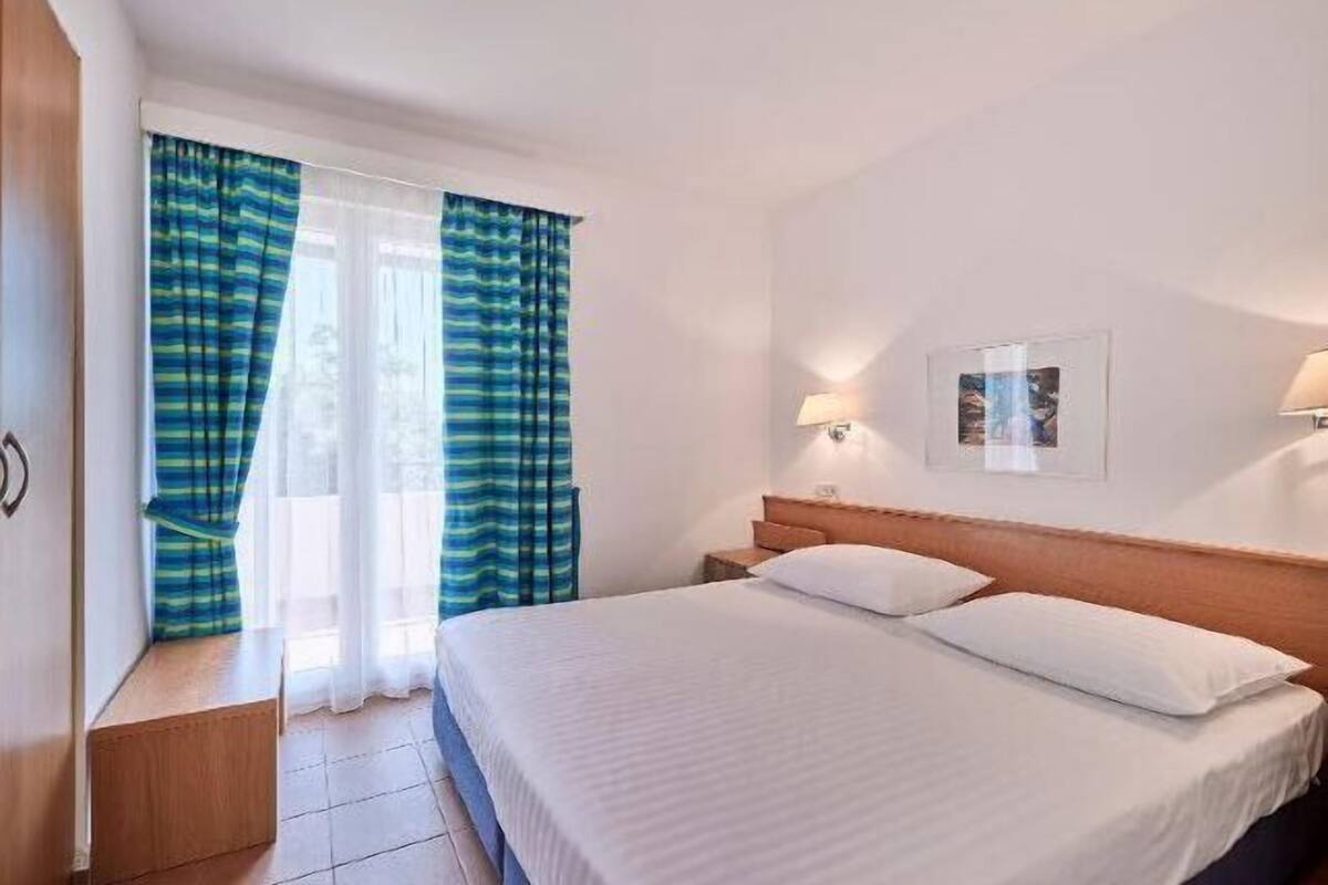 Una habitación de hotel con una cama, cortinas, una mesita de noche, una lámpara de pared y un cuadro enmarcado.