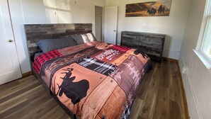 A bedroom with a wooden headboard, a bed with a patterned blanket, a ceiling fan, a door, and a painting on the wall. - Base Camp Retreat (Glendo)