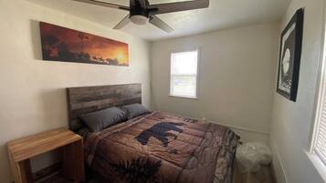 A bedroom with a bed, a nightstand, a ceiling fan, a window, and a painting on the wall.