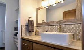 Un bagno con un lavandino bianco, uno specchio, un portascopevoli e un armadietto in legno.