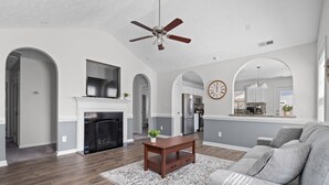 A spacious living room with a fireplace, a ceiling fan, and a large window. - Quaint Cottage-3BR/2B House with Arcade and Fire Pit in Quiet Part of Hope Mills (Hope Mills)
