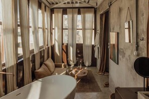 A room with a large bathtub, a sofa, a small table, and curtains on the windows. - Mona Athens (Athens)