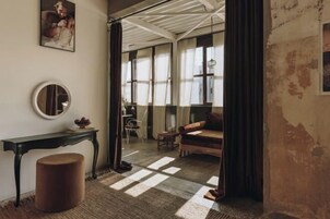 A cozy room with a round mirror, a small table, a cushioned bench, and a view through a window with sheer curtains.