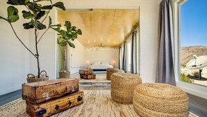 Un dormitorio acogedor con un gran ventanal, una cama y muebles rústicos, incluyendo un baúl y otomanas.
