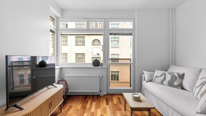 Een moderne woonkamer met een bank, televisie, houten dressoir en een uitzicht op gebouwen door grote ramen.