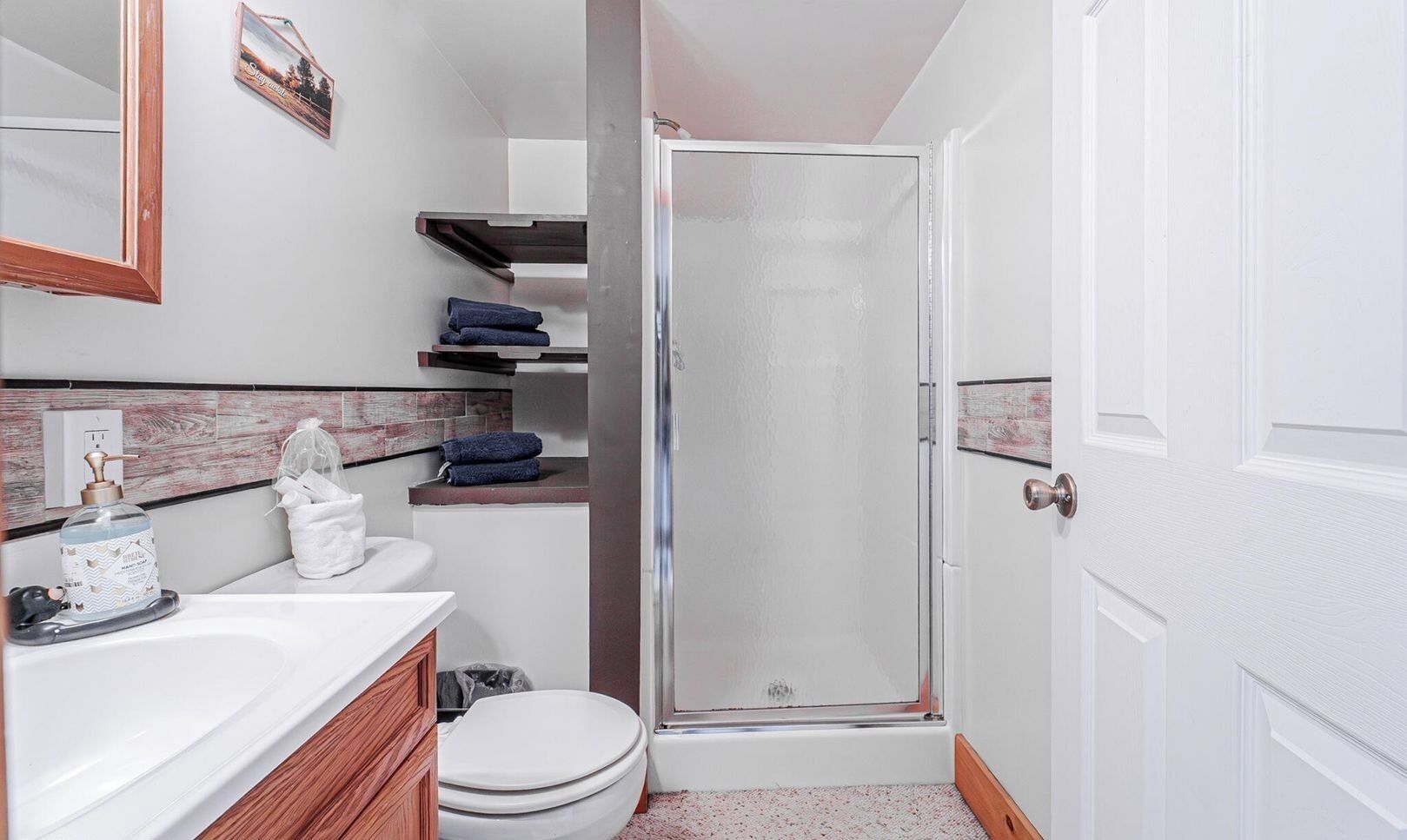 Un bagno con lavandino, water e doccia bianchi. Ci sono scaffali sopra il water e uno specchio sopra il lavandino.