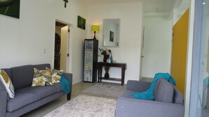 Ein Wohnzimmer mit grauem Sofa, einem kleinen grauen Sessel, einem Couchtisch und einer Lampe. An der Wand hängt ein gerahmtes Bild und ein kleines Regal mit Dekoration.