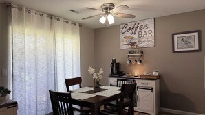 A dining area with a table set for four, a ceiling fan, a coffee bar, and a framed picture on the wall.