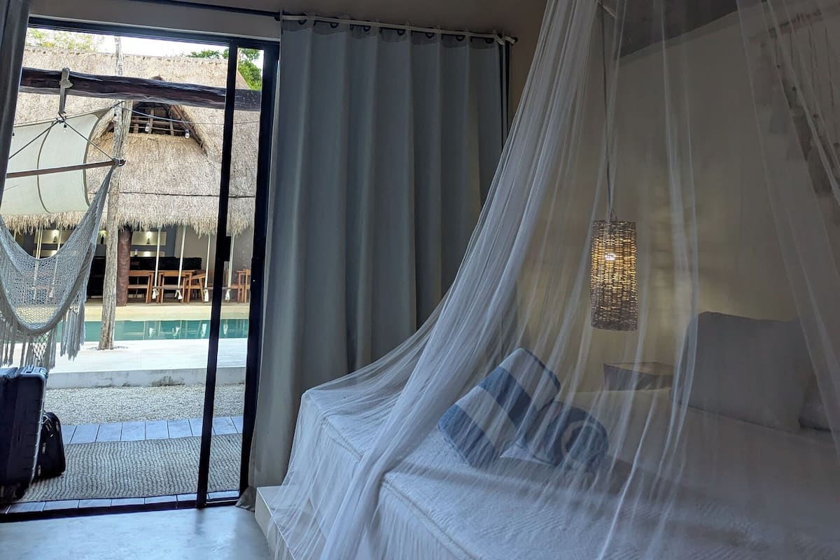 A room with a bed under a mosquito net, a ceiling fan, and a view of an outdoor area with a thatched roof.