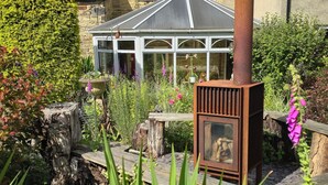 Una chimenea rústica, un edificio de piedra con tejado de baldosas y un jardín con diversas plantas y un banco de madera.