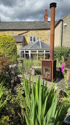 A rustic chimney, a stone building with a tiled roof, and a garden with various plants and a wooden bench. - 17th Century Farmhouse (Tibshelf)