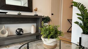 Un interno moderno con un tavolo con piano in vetro, vasi decorativi, un cesto di vimini con verde e uno specchio sopra una mensola scura.