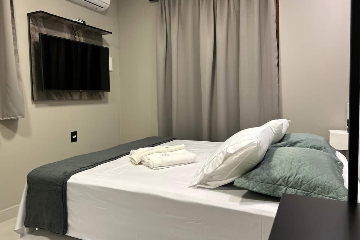 A neatly made bed with white and green pillows, a grey blanket, and folded towels on a clean white mattress.