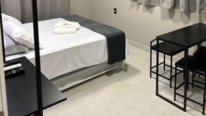 A compact hotel room with a bed, a small table, a TV mounted on the wall, and an air conditioning unit. - Apartamentos Bora Bora Hospedagens (Joinville)