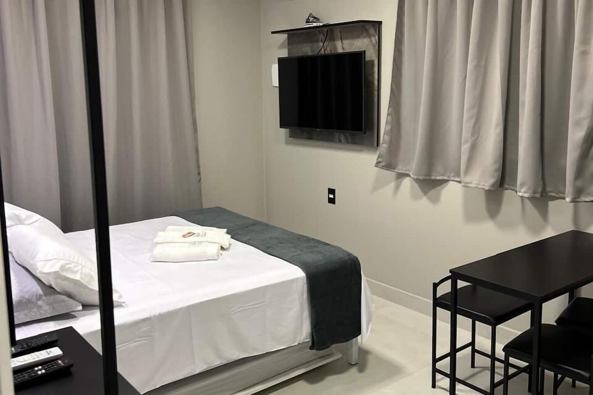 A compact hotel room with a bed, a small table, a TV mounted on the wall, and an air conditioning unit.