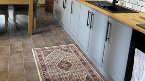 A modern kitchen with light-colored cabinets, a wooden countertop, and a built-in oven. There is a dining table with chairs and a rug on the floor.