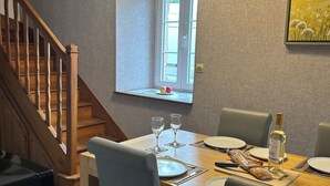 A dining area with a wooden table set for two, a bottle of wine, and a window with a view of a field.