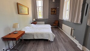 Un dormitorio con cama, mesita de noche de madera, una silla y dos ventanas con cortinas.