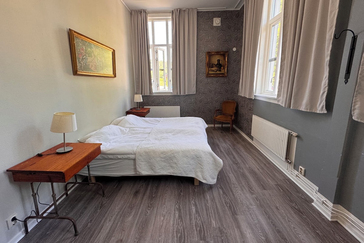 Een slaapkamer met een bed, een houten nachtkastje, een stoel en twee ramen met gordijnen.